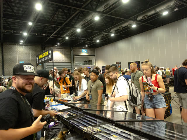 A smiling trader behind a glass counter while a crowd works along the tables behind him.