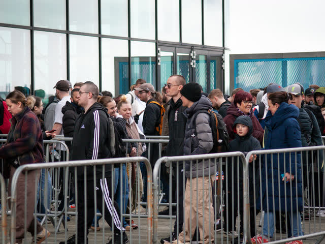 Collectors queuing behind metal barriers at the entrance, bright sky through the windows behind.