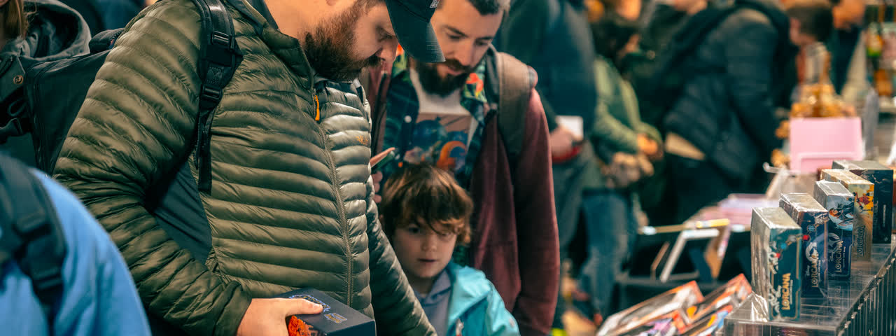 A man holding a sealed box at a busy stand, a child beside him looking over the table.